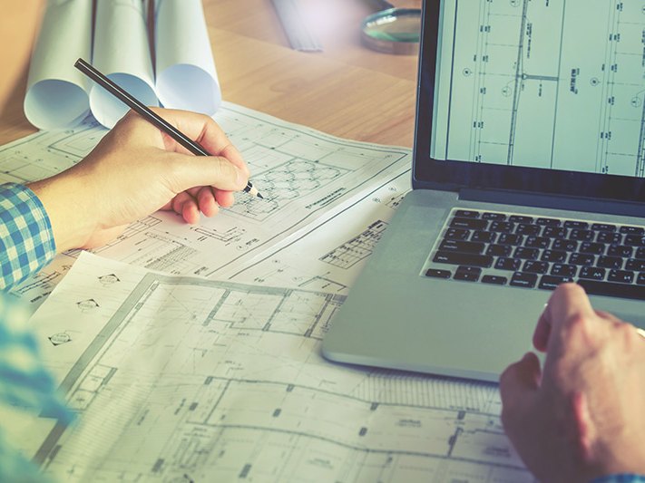 Mani che tracciano uno schema tecnico su fogli di progetto accanto a un computer portatile